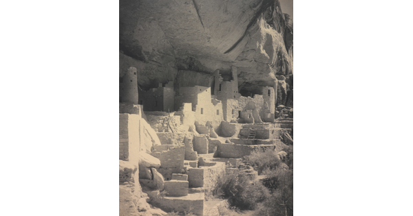 A black-and-white photograph of an abandoned, crumbling rock city built into the side of a desert cliff.