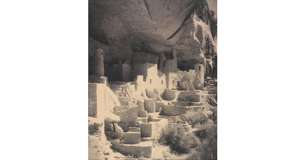 A black-and-white photograph of an abandoned, crumbling rock city built into the side of a desert cliff.