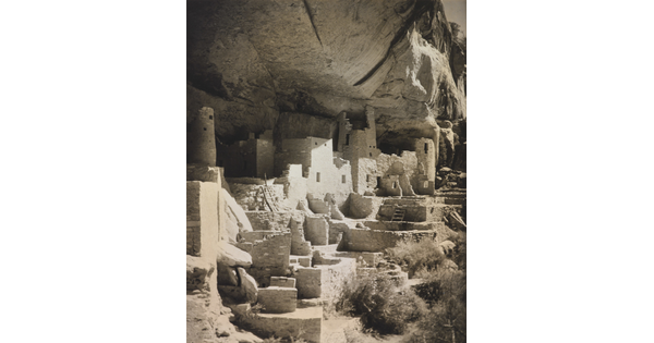 A black-and-white photograph of an abandoned, crumbling rock city built into the side of a desert cliff.