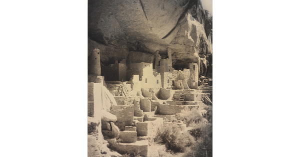 A black-and-white photograph of an abandoned, crumbling rock city built into the side of a desert cliff.