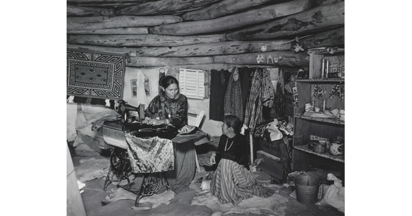 A black-and-white photograph of two Indigenous women seated, talking, and sewing inside of a low-ceilinged, wood structure.
