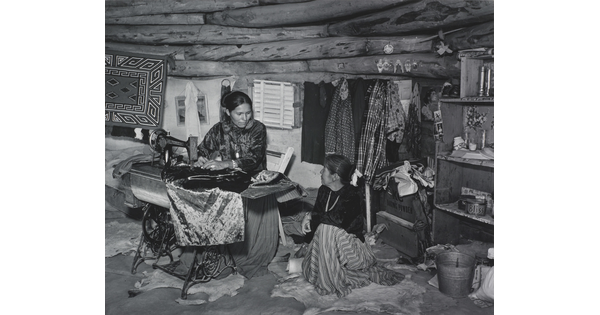 A black-and-white photograph of two Indigenous women seated, talking, and sewing inside of a low-ceilinged, wood structure.