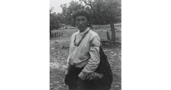 A black-and-white photograph of an Indigenous man carrying a large bag.