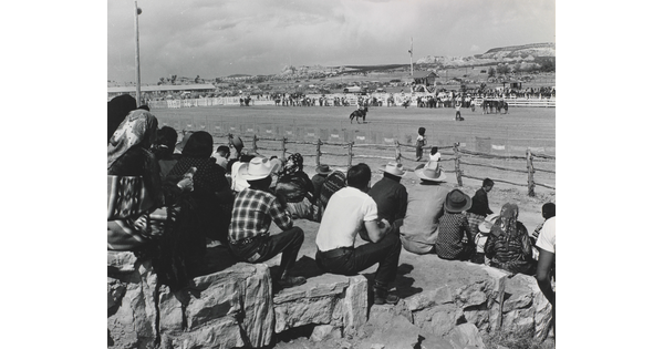 A black-and-white photograph of a group of spectators watching cowboys and horses in an outdoor arena.