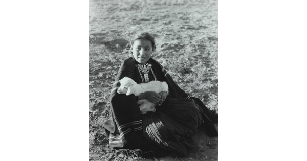 A black-and-white photograph of an Indigenous girl sitting on the ground while holding a lamb.