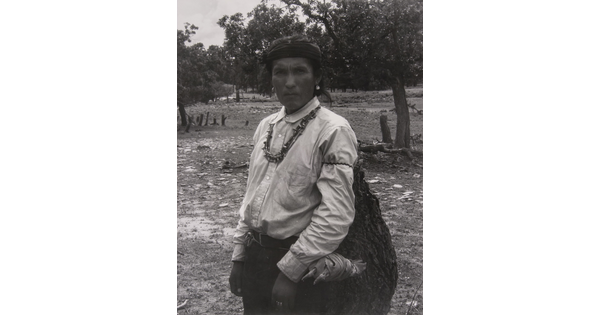 A black-and-white photograph of an Indigenous man carrying a large bag.