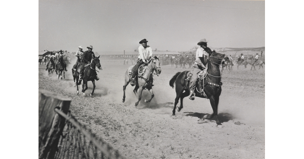 A black-and-white photograph of men wearing cowboy hats riding running horses on a dusty track.