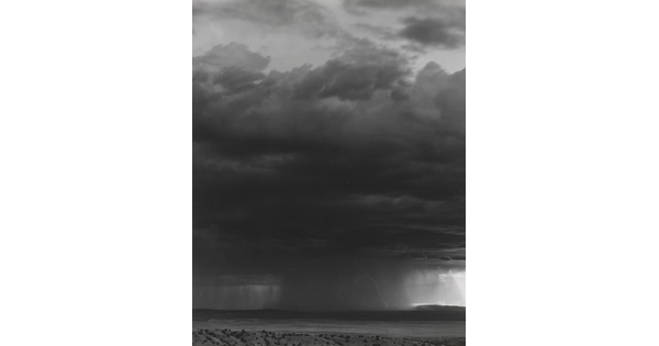 A black-and-white photograph of a dark storm cloud, heavy rain, and lightening over a desert.