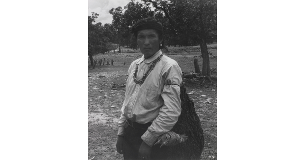 A black-and-white photograph of an Indigenous man carrying a large bag.