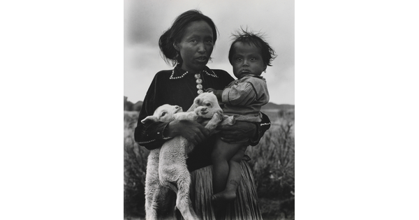 A black-and-white portrait photograph of a Native American woman holding a toddler on one hip and two lambs in her other arm.
