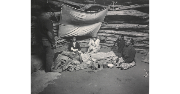 A black-and-white photograph of an Indigenous man laying on a pallet surrounded by other Indigenous people as a White woman holds a glass jar out to him.