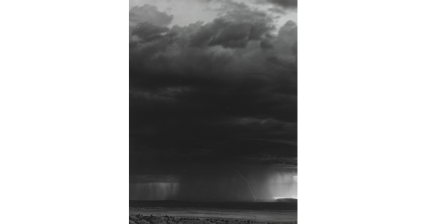 A black-and-white photograph of a dark storm cloud, heavy rain, and lightening over a desert.