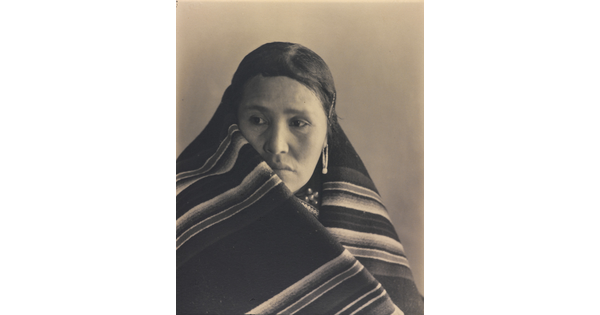 A black-and-white portrait photograph of an Indigenous woman with a blanket wrapped around her neck and shoulders.