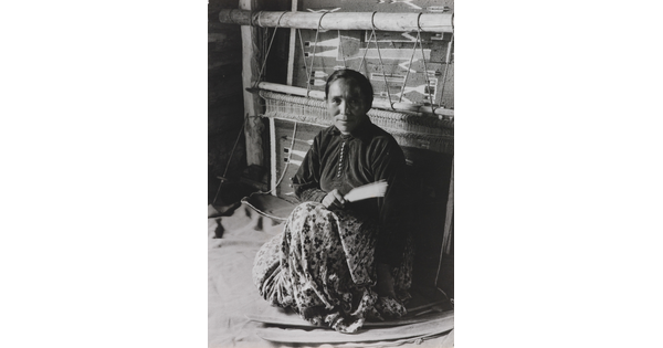 A black-and-white photographic portrait of a Native American woman sitting on the floor in front of a large loom.