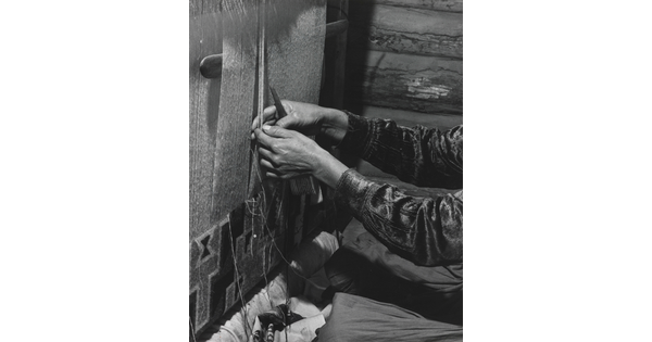 A black-and-white photograph of a person's hands weaving a design into a tapestry on a loom.