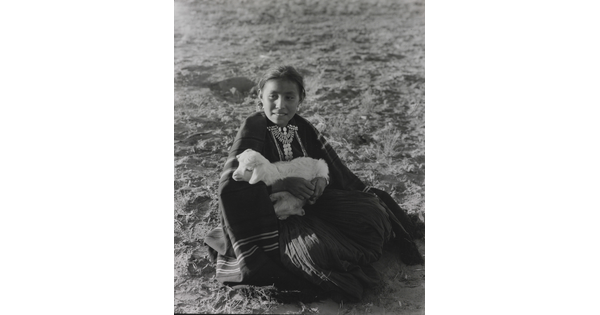 A black-and-white photograph of an Indigenous girl sitting on the ground while holding a lamb.