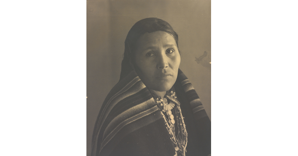 A black-and-white portrait photograph of an Indigenous woman with a blanket wrapped around her neck and shoulders.
