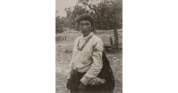 A sepia-toned photograph of an Indigenous man carrying a large bag.