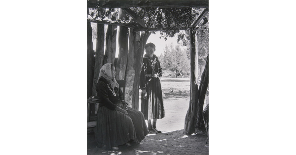 A black-and-white photograph of two Indigenous women, one seated and one standing, in a wooden structure open to outside.