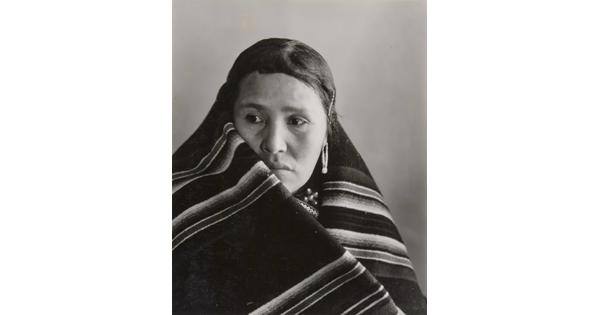 A black-and-white portrait photograph of an Indigenous woman with a blanket wrapped around her neck and shoulders.