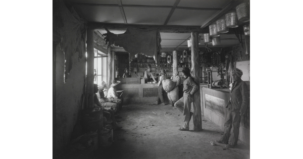 A black-and-white photograph of Indigenous people in a shop with a variety of products such as animal hides, tin pails, and lanterns hanging from the ceiling.