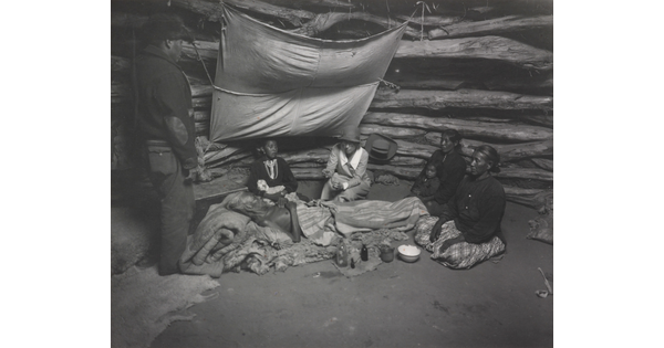 A black-and-white photograph of an Indigenous man laying on a pallet surrounded by other Indigenous people as a White woman holds a glass jar out to him.