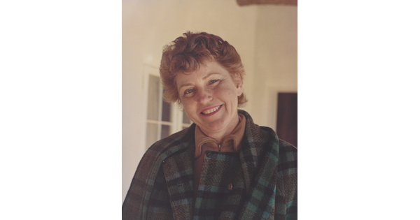 A color portrait photograph of a smiling, fair-skinned woman with red hair, wearing a plaid jacket, and standing on a porch.