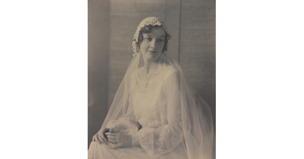 A black-and-white portrait photograph of a seated young White woman, hands folded on her lap, wearing a wedding dress with a long veil.