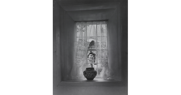 A black-and-white photograph of a sheer curtained window with a Native American-style pot on the sill as a White woman cleans the window from outside.