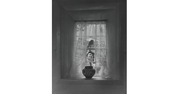 A black-and-white photograph of a sheer curtained window with a Native American-style pot on the sill as a White woman cleans the window from outside.