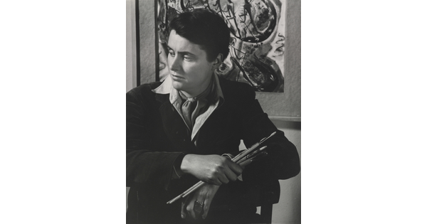 A black-and-white portrait photograph of a seated White woman holding paintbrushes in front of an abstract painting.