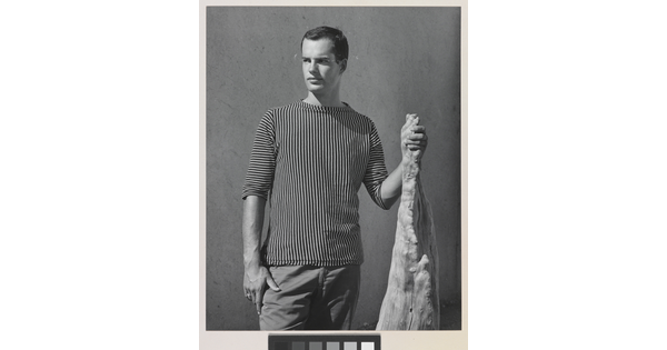 A black-and-white photograph of a White man with wearing a shirt with vertical stripes and holding a wooden object.