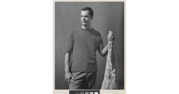 A black-and-white photograph of a White man with wearing a shirt with vertical stripes and holding a wooden object.