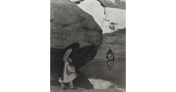 A black-and-white photograph of a woman with a long headscarf standing at the edge of a water hole surrounded by boulders and rock walls.