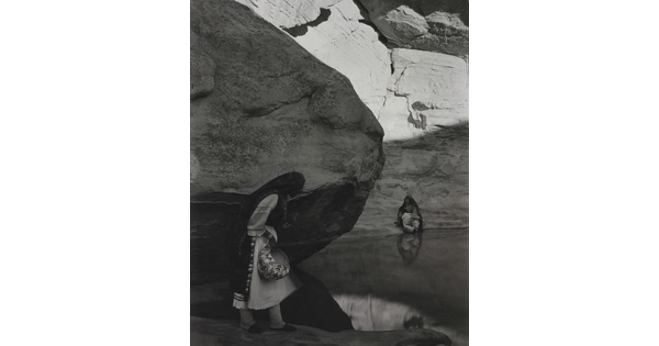A black-and-white photograph of a woman with a long headscarf standing at the edge of a water hole surrounded by boulders and rock walls.