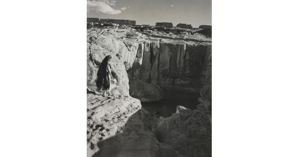 A black-and-white photograph of a woman with a long headscarf on a cliff, overlooking a water hole at the bottom.