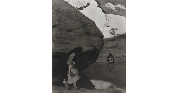 A black-and-white photograph of a woman with a long headscarf standing at the edge of a water hole surrounded by boulders and rock walls.