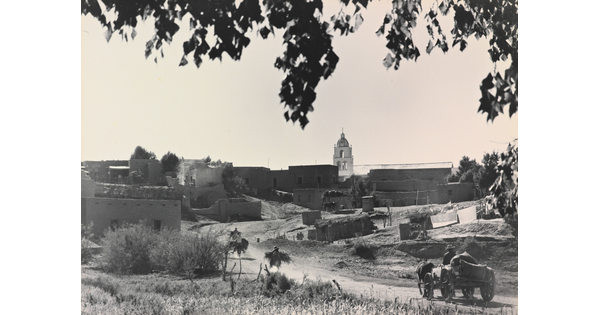 A black-and-white photograph of a town with adobe buildings and wagons and horses traveling on a dusty road.