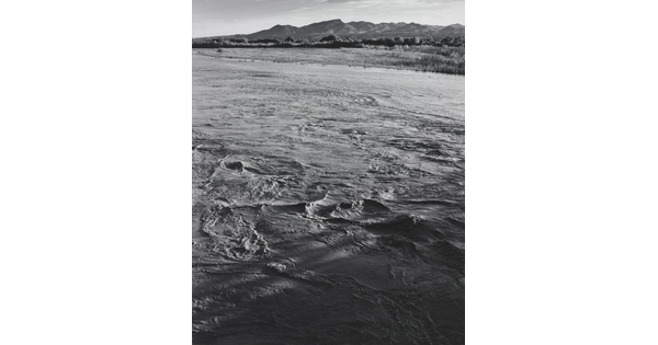 A black-and-white photograph of a choppy river and then grassy plains and mountains beyond the shore.