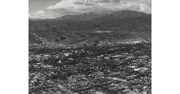 A black-and-white aerial photograph of a town in a mountain valley beneath puffy white clouds.