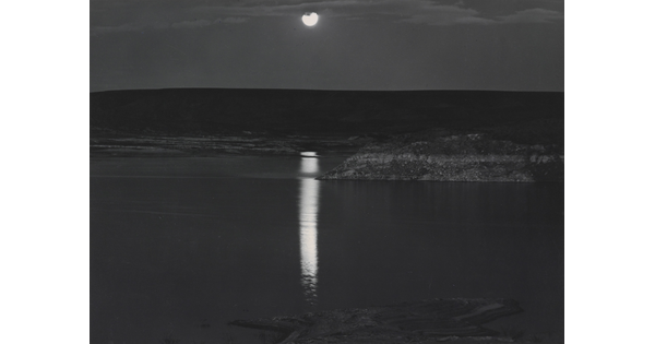 A black-and-white photograph of a night sky and full moon reflected across calm water.