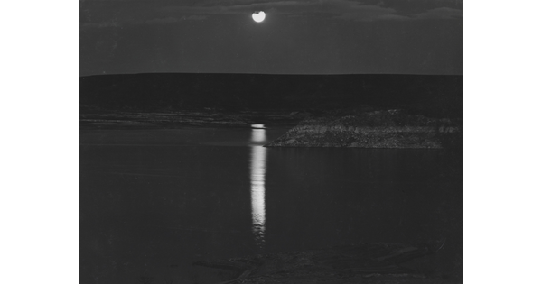 A black-and-white photograph of a night sky and full moon reflected across calm water.