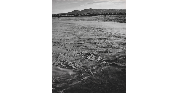 A black-and-white photograph of a choppy river and then grassy plains and mountains beyond the shore.