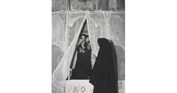 A black-and-white photograph of a woman wearing a black shawl over her head facing a statue of a saint holding a large key.