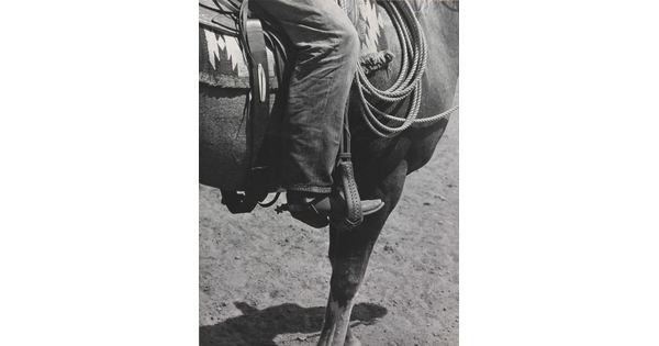 A black-and-white photograph of a person's leg in a cowboy boot, including spurs, sitting astride a horse, viewing only the horse's front quarter.