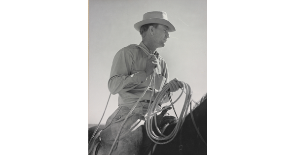 A black-and-white photograph of a White man wearing a brimmed hat holding a lasso astride a dark horse.