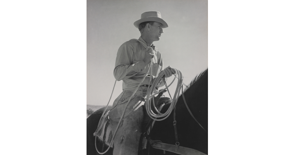 A black-and-white photograph of a White man wearing a brimmed hat holding a lasso astride a dark horse.