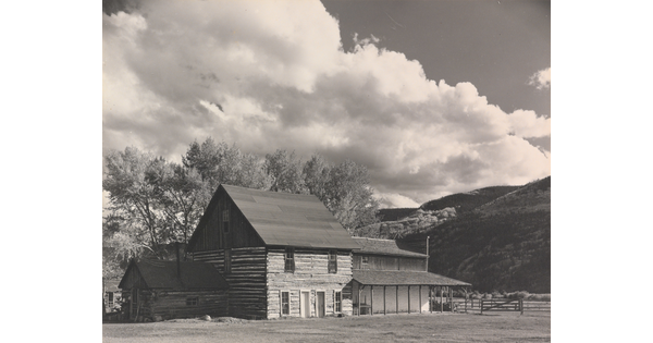 A black-and-white photograph of a three-story log building with mountains and large puffy clouds in the background.