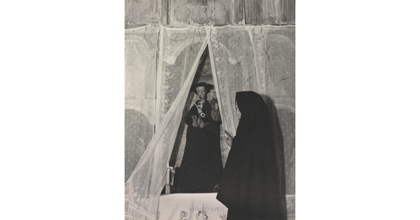 A black-and-white photograph of a woman wearing a black shawl over her head facing a statue of a saint holding a large key.