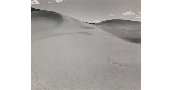 A black-and-white photograph of a rippling sand dune rising up into a sky dotted with a few small clouds.